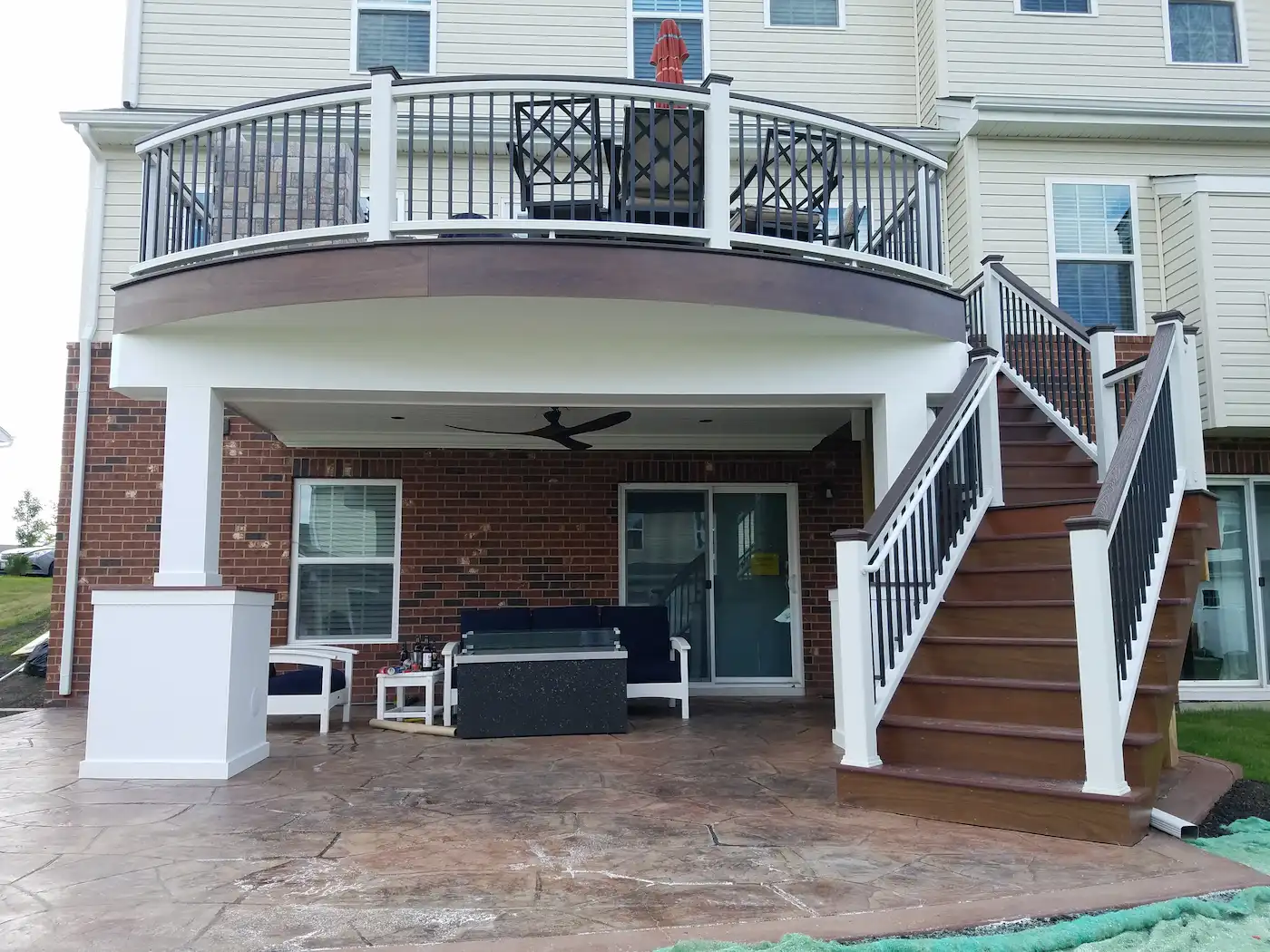 A raised, slightly curved Trex composite deck with an angled staircase, built over a finished Trex RainEscape under-deck living space featuring a ceiling fan and outdoor furniture.